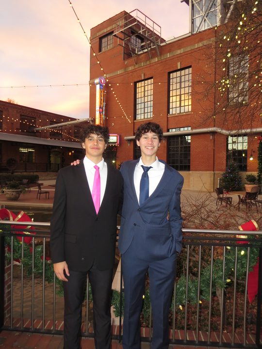 Ayden and Alex in suits, standing in front of a brick building strung with lights.