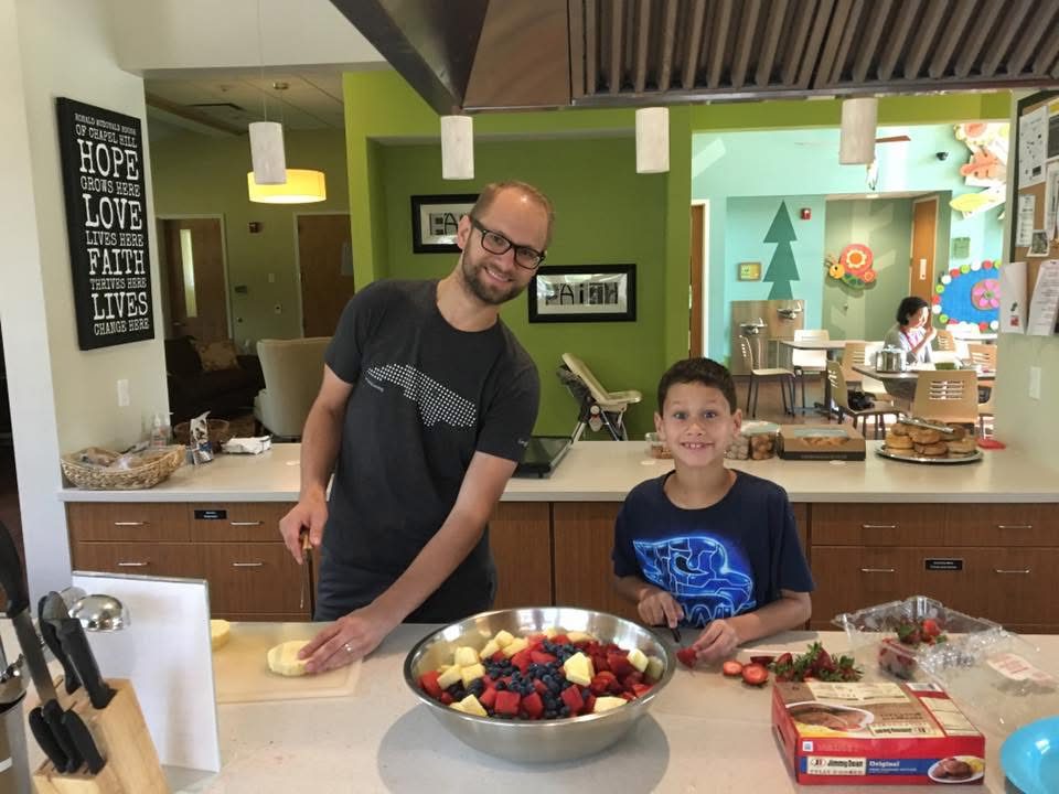 A young Ayden smiling while cutting strawberries in the kitchen at the Ronald McDonald House, next to an adult volunteer.